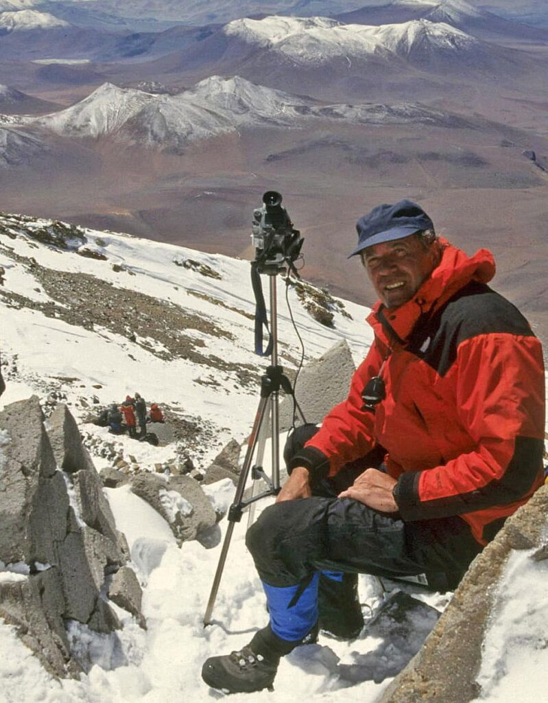 Archaeologist Johan Reinhard at the Summit of Llullaillaco Volcano During the Discovery of the Inca Mummies, 1999 | Peru Travel Blog Archaeologist Johan Reinhard in a red high-altitude jacket sits beside a camera on a tripod at the snow-covered summit of Llullaillaco volcano at 6,739 meters, with team members visible below on the slope and the vast Atacama Puna landscape stretching to the horizon, Argentina-Chile border, 1999