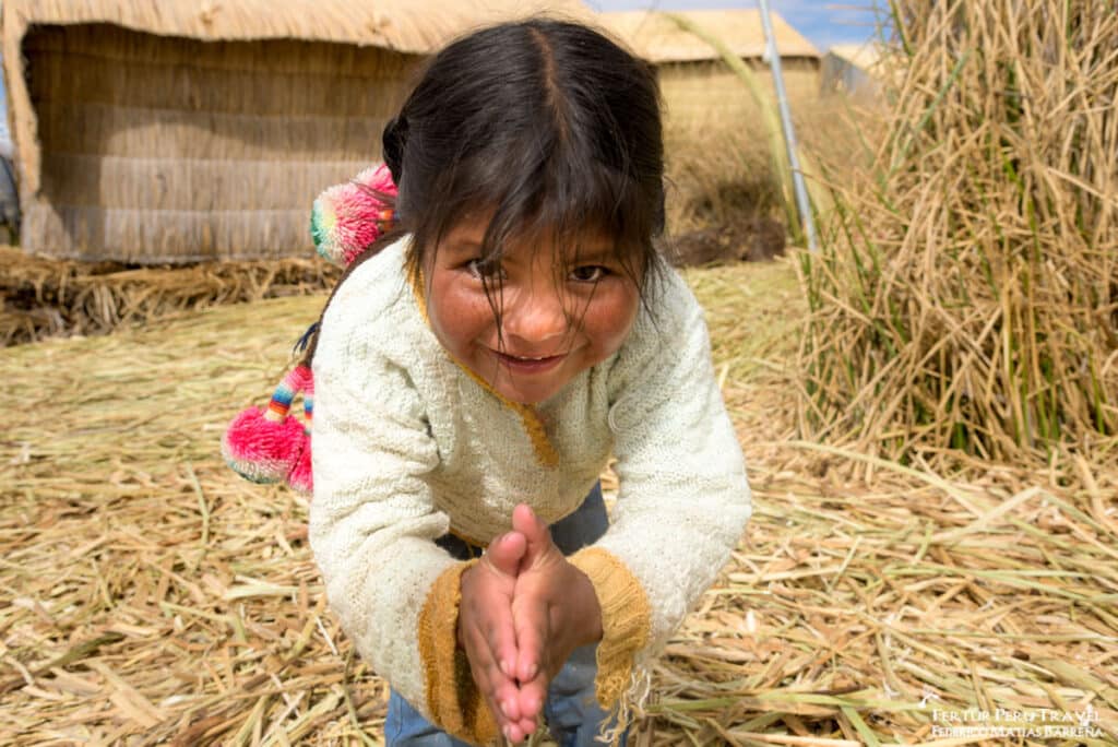 Uros Girl Reaching Out in Welcome on the Floating Reed Islands of Lake Titicaca, Puno, Peru | Peru Travel Blog A disarming and joyful close-up portrait of a young Uros girl on the floating islands of Lake Titicaca, leaning forward with both hands stretched out toward the camera in a gesture of playful greeting or offering.