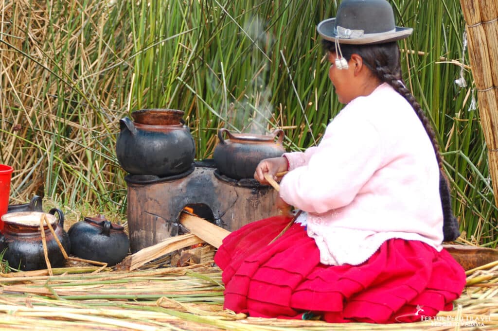Uros Woman Cooking Over a Wood Fire on a Totora Reed Floating Island | Peru Travel Blog An Uros woman in a red skirt, pink sweater, and grey bowler hat tends a wood fire under clay cooking pots on a traditional clay stove on the totora reed surface of a floating island, surrounded by standing totora reeds, Lake Titicaca, Puno, Peru