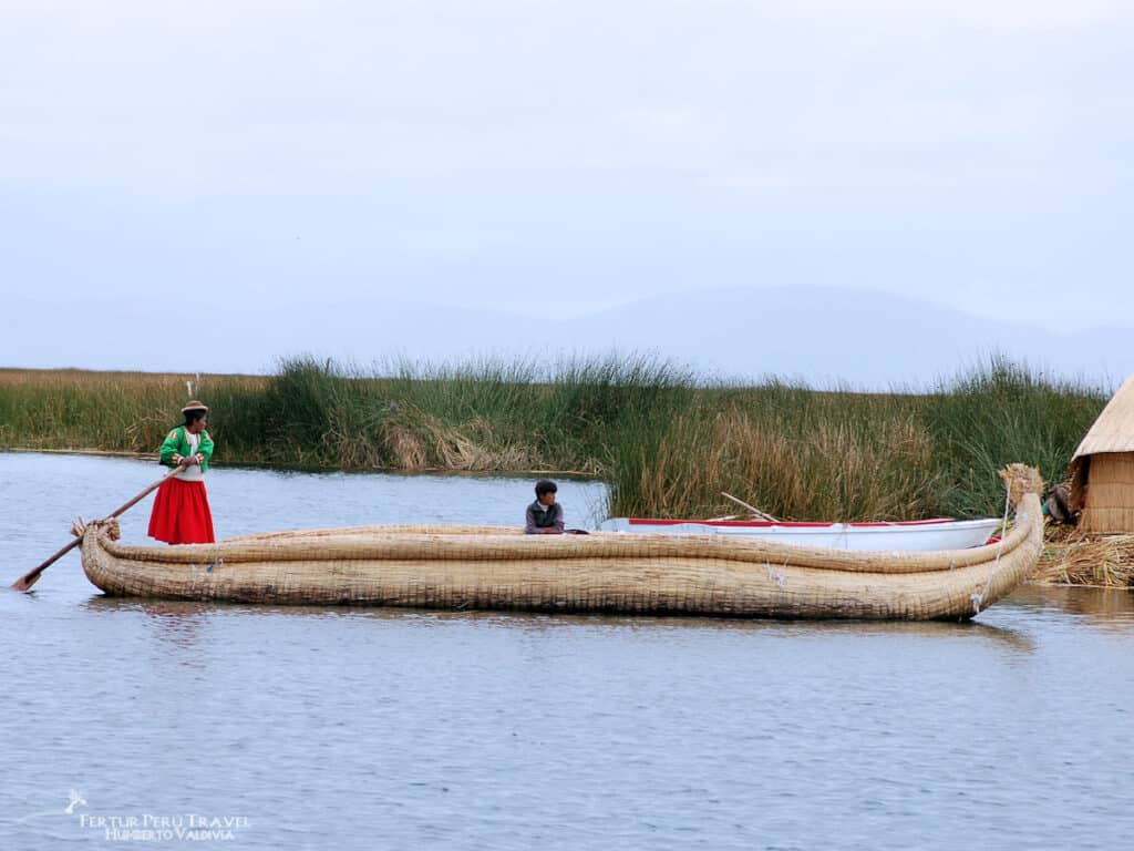 Uros Woman Paddling a Traditional Totora Reed Boat on Lake Titicaca | Peru Travel Blog Uros woman in a red skirt and green jacket stands at the stern paddling a large traditional totora reed boat with a child seated inside, surrounded by totora reed beds on the calm waters of Lake Titicaca, Puno, Peru