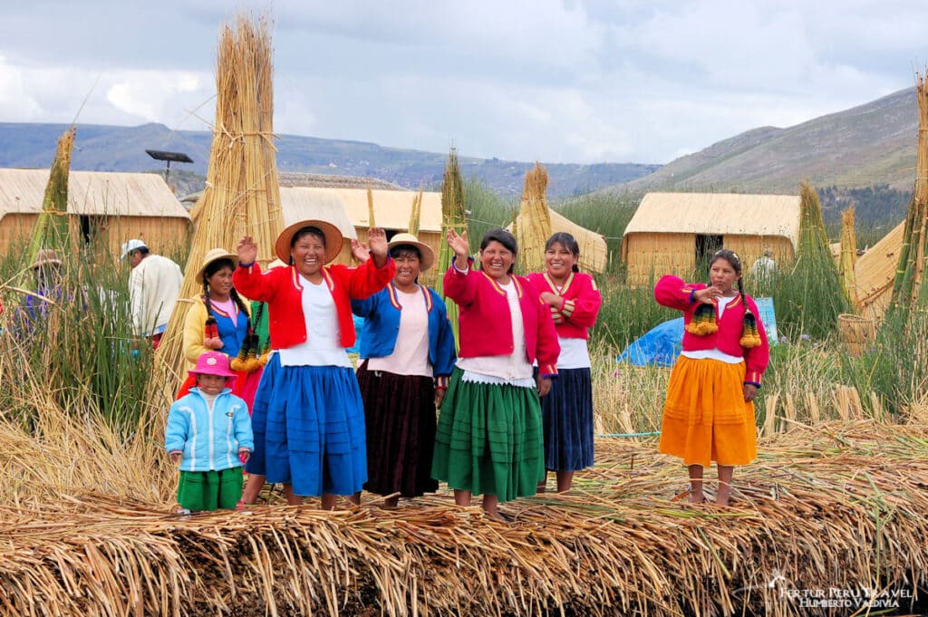 Uros Women and Children Waving Goodbye on the Floating Reed Islands of Lake Titicaca | Peru Travel Blog A group of smiling Uros women and a small child in vibrant traditional dress wave farewell to departing visitors on the totora reed surface of a floating island, with traditional reed dwellings, bundled totora stalks, and the hills above Puno visible behind them, Lake Titicaca, Peru