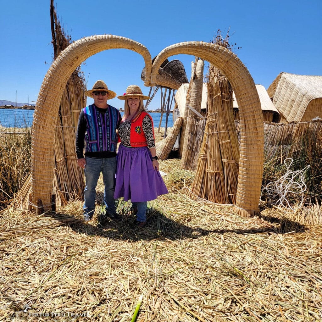 Fertur Peru Travel Clients Visiting the Uros Floating Islands, Lake Titicaca, Peru | Peru Travel Blog A smiling couple dressed in borrowed traditional Uros clothing and straw hats pose beneath a decorative totora reed arch on the Uros floating islands on Lake Titicaca, with the deep blue lake and clear Andean sky visible behind them, Puno, Peru