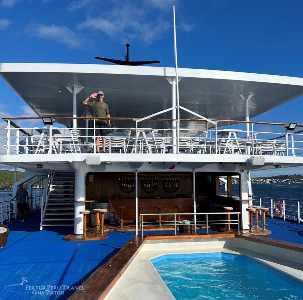 Galápagos Legend Cruise Ship Pool Deck, Galápagos Islands | Photo: Gina Turton | Peru Travel Blog A traveler on Galapagos Islands cruise waves from the upper observation deck of the Galápagos Legend luxury cruise yacht, with a sparkling pool, open-air bar, and polished teak decking in the foreground, and a green island shoreline visible across the water in the background. Photo credit: Fertur Perú Travel / Gina Turton.
