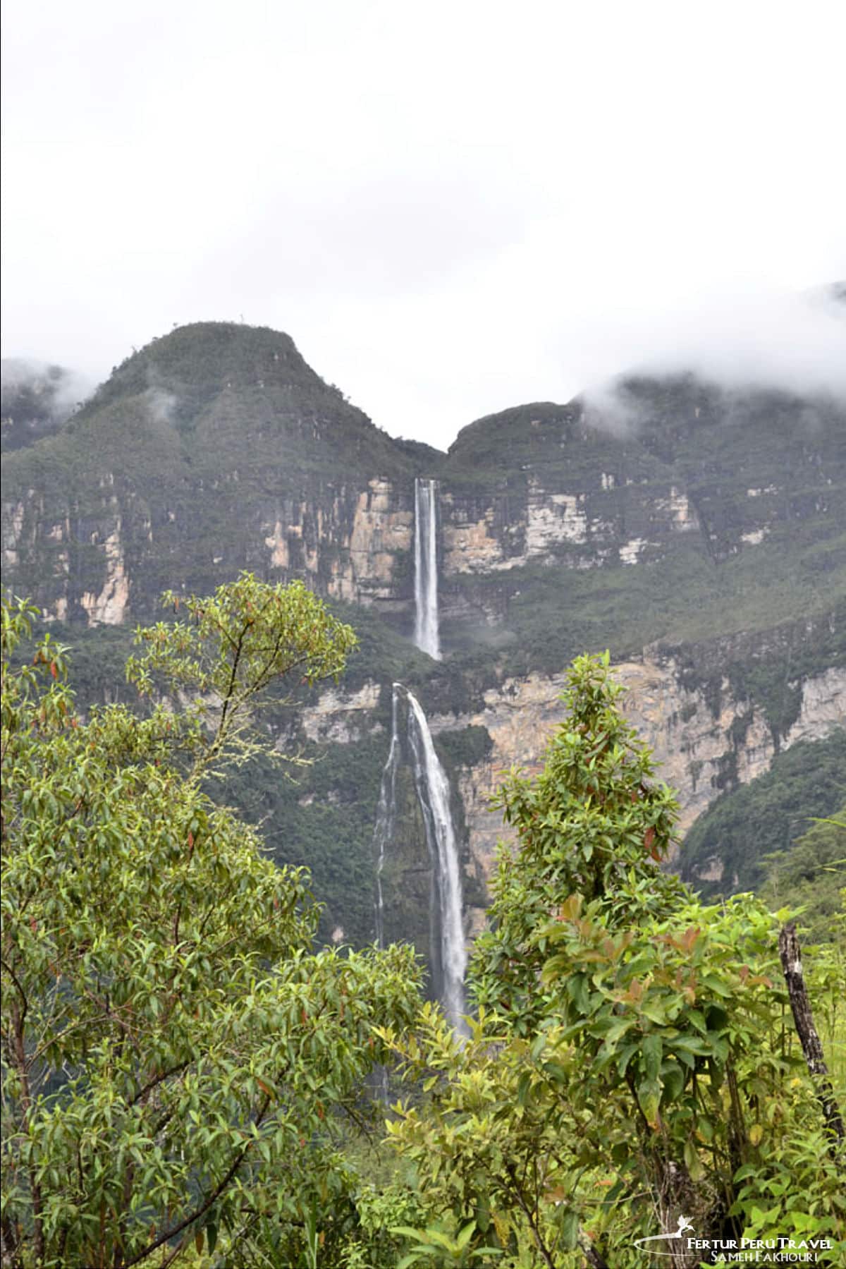 Gocta Waterfall in the Cloud Forest, Amazonas, Peru | Photo: Sameh Fakhouri | Peru Travel Blog A distant view of Gocta Falls, a dramatic two-tiered waterfall cascading down a sheer cliff face in the cloud forest of Amazonas, Peru, framed by lush tropical vegetation and low-hanging mist. Photo credit: Sameh Fakhouri.