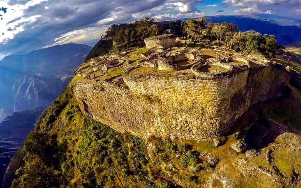 Aerial View of Kuélap Fortress, Amazonas, Peru | Peru Travel Blog A sweeping aerial view of the ancient Kuélap fortress perched on a dramatic ridgeline in the Amazonas region of Peru, revealing its massive stone exterior walls, circular interior structures, and the deep valley far below. Photo credit: Heinz Plenge.