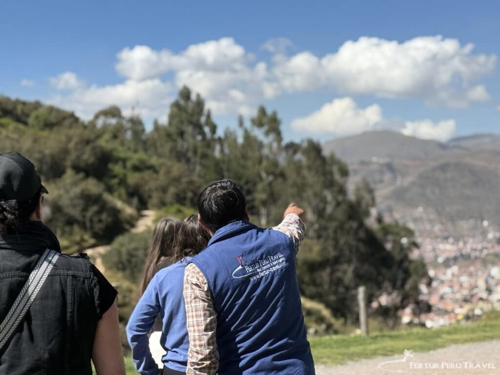 Fertur Travel Guide Overlooking Cusco from Sacsayhuamán | Peru Travel Blog A Fertur Peru Travel guide, wearing a branded blue vest, points out landmarks across the Cusco valley to a small group of travelers from a hillside viewpoint near Sacsayhuamán. Photo credit: Fertur Perú Travel.