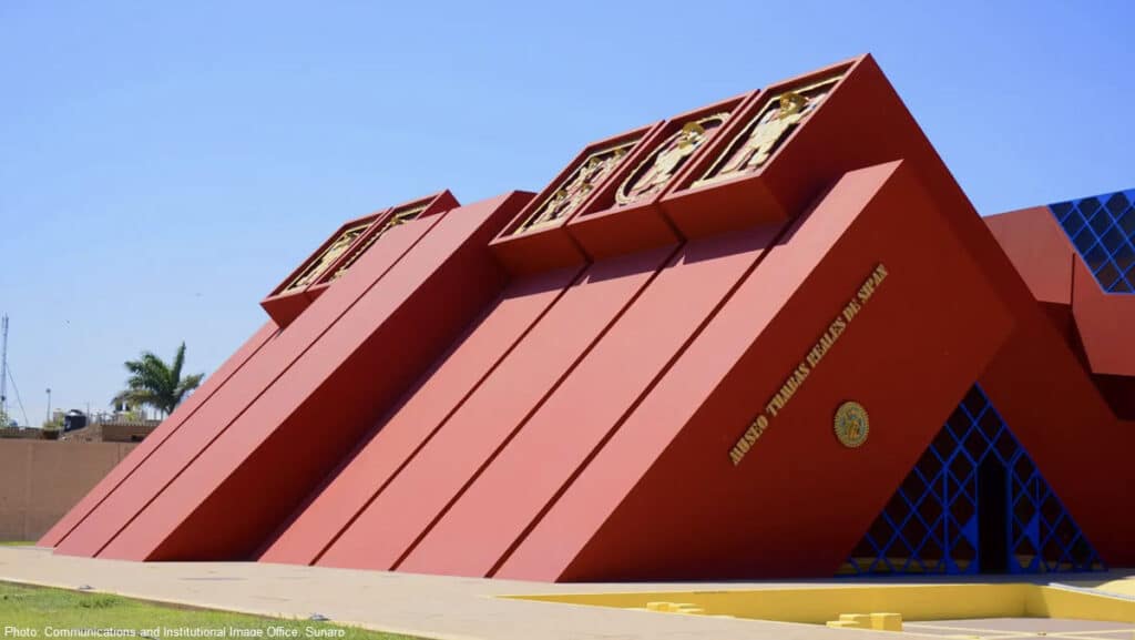 Museo Tumbas Reales de Sipán, Lambayeque, Peru | Peru Travel Blog The striking stepped-pyramid exterior of the Museo Tumbas Reales de Sipán in Lambayeque, Peru, its bold terracotta-red facade rising at an angle with golden figural reliefs set into recessed panels along the roofline and blue geometric lattice framing the entrance. Photo: Communications and Institutional Image Office, Sunarp.