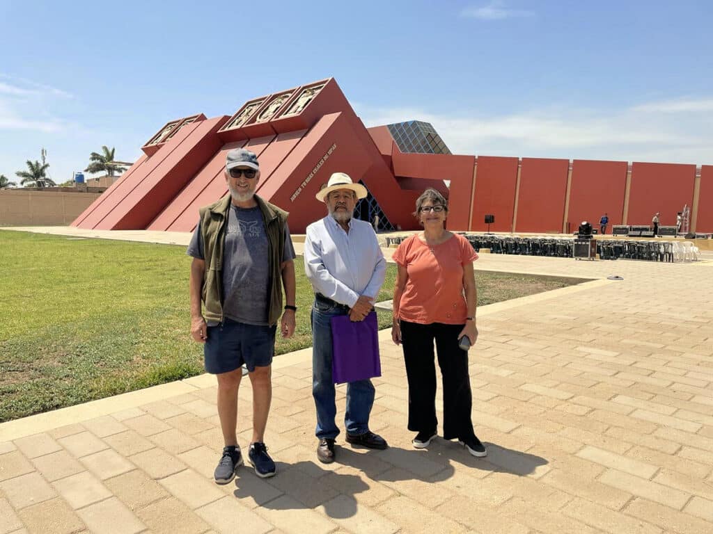 Fertur Travelers Meet Archaeologist Walter Alva (center) at Museo Tumbas Reales de Sipán | Peru Travel Blog Two Fertur Travel clients pose with Peruvian archaeologist Walter Alva (center, in white shirt and panama hat) in front of the terracotta-red stepped-pyramid facade of the Museo Tumbas Reales de Sipán in Lambayeque, Peru. Photo credit: Fertur Perú Travel.