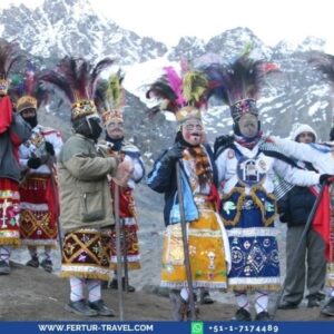 Group of dancers in elaborate traditional Andean ceremonial costumes with colorful feathered headdresses and ornately embroidered tunics, standing in front of snow-capped mountains during the Qoyllur Rit'i festival in Peru