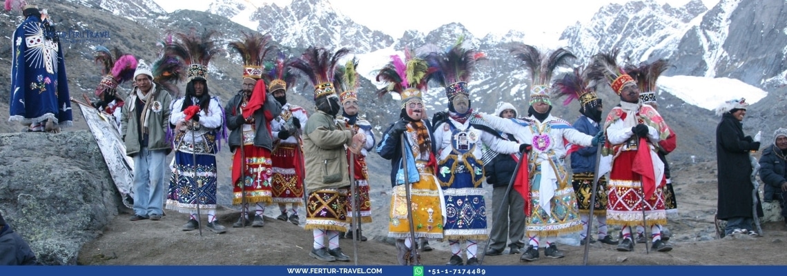 Group of dancers in elaborate traditional Andean ceremonial costumes with colorful feathered headdresses and ornately embroidered tunics, standing in front of snow-capped mountains during the Qoyllur Rit'i festival in Peru
