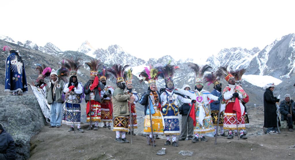 Qoyllur Rit’i pilgrimage dancers in ornate Andean costumes and feathered headdresses standing on a rocky mountainside beneath snowcapped peaks in Peru.