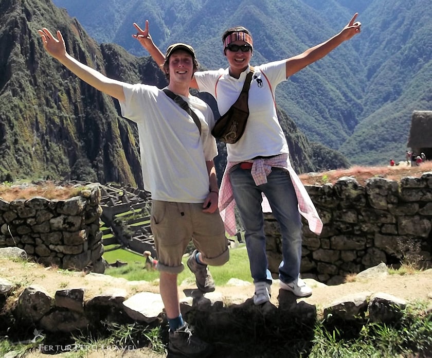 Dos viajeros felices en Machu Picchu tras una caminata con Fertur Peru Travel, con los brazos en alto en señal de celebración y haciendo el gesto de la paz.