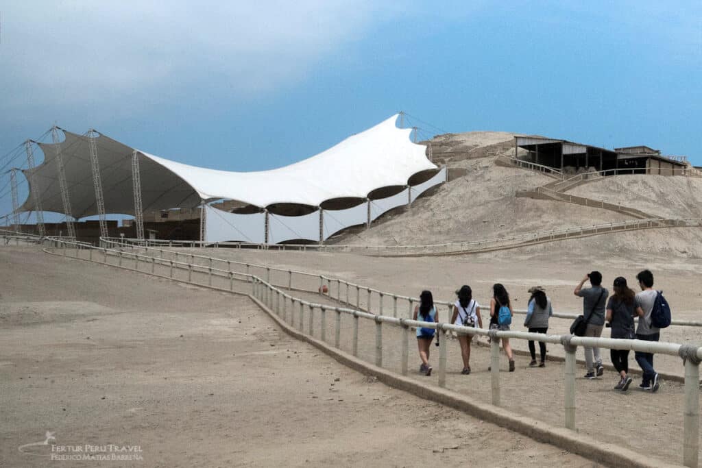 Visitors walk a guided path toward the ancient adobe pyramid of Huaca Cao Viejo, protected by a modern tensile roof structure at the El Brujo archaeological complex near Trujillo, Peru.