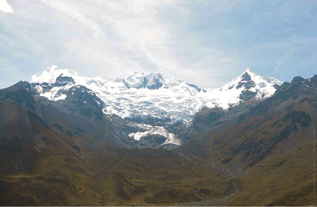 Nevado Huaytapallana, a snow-capped mountain range in the Junín Region of Peru, rising above a sweeping high-altitude valley covered in golden-brown grasslands, with glaciers cascading down its rocky slopes beneath a partly cloudy sky.